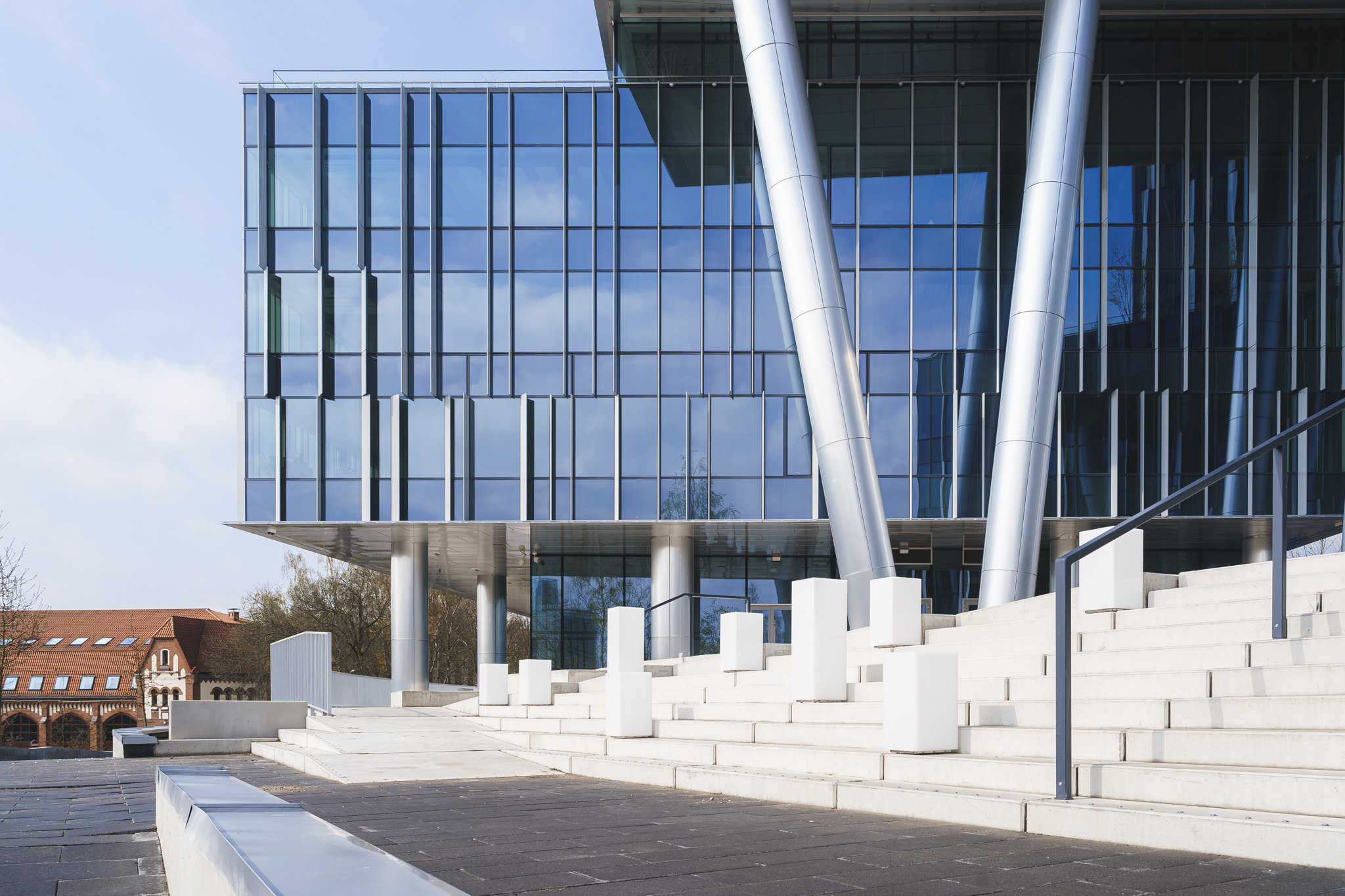 Low-angle view of a modern glass office building with white exterior stairs and silver support columns under a blue sky.